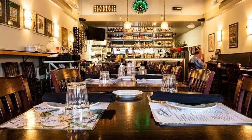 Restaurant interior with set tables, wooden chairs, a bar in the background, and a man dining.