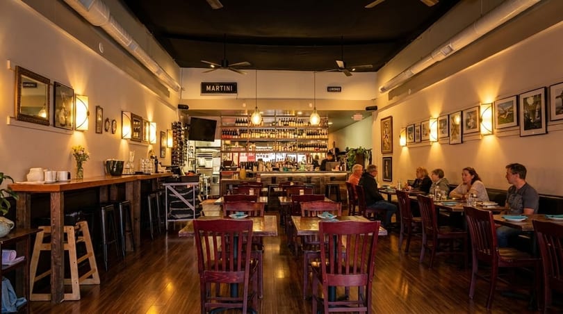 Restaurant interior with patrons dining, a long bar, and a wall with framed art and sconces.